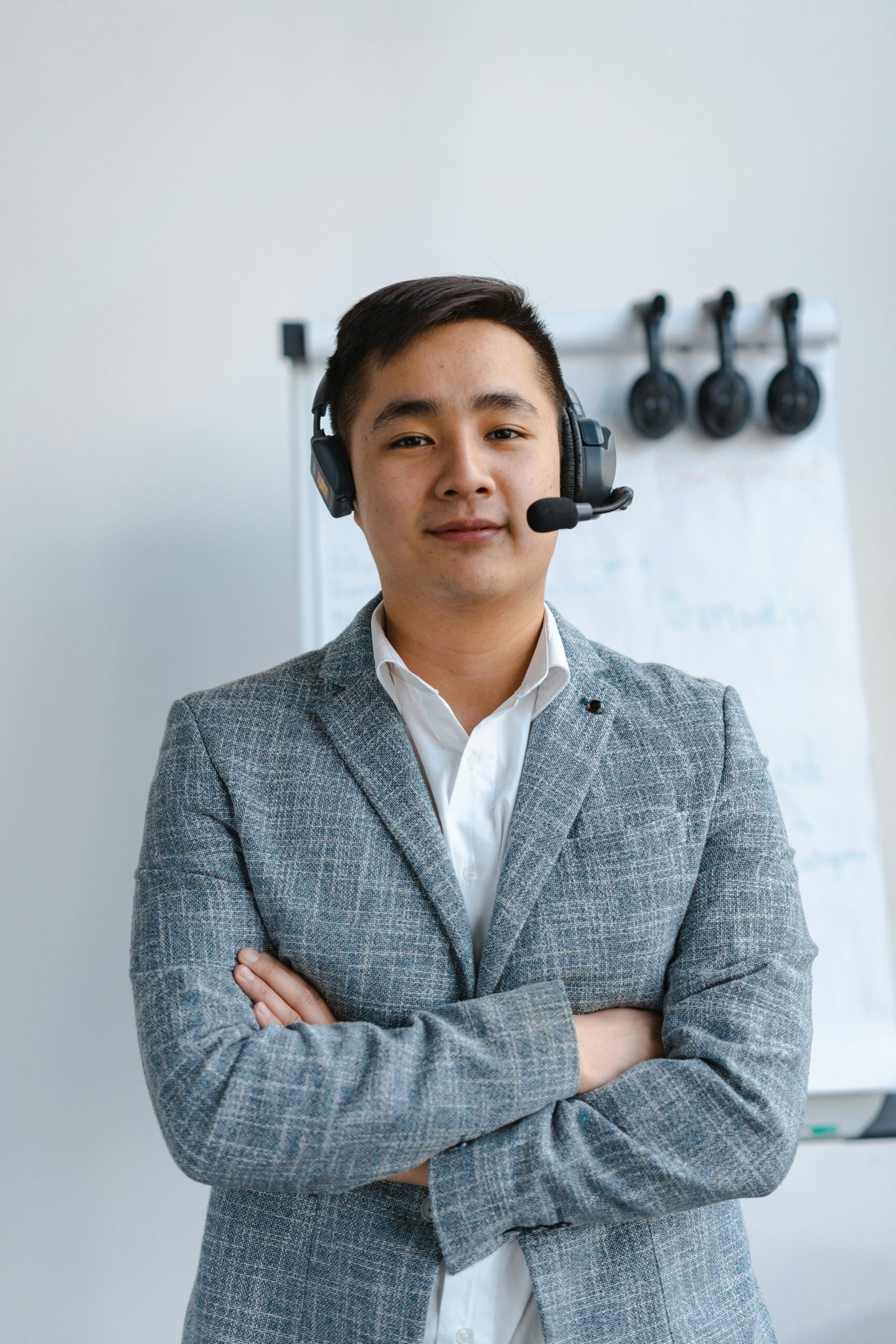 Asian call center agent wearing a headset and gray suit standing confidently indoors.